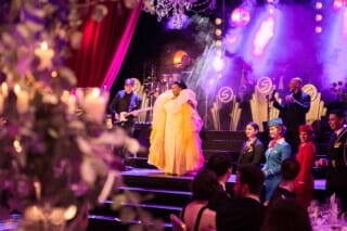 Stage performance by a singer in a yellow dress, surrounded by musicians and an elegant audience.
