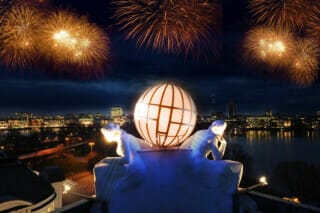 Feuerwerk über der Stadt mit leuchtender Skulptur und Nachtpanorama am Wasser.