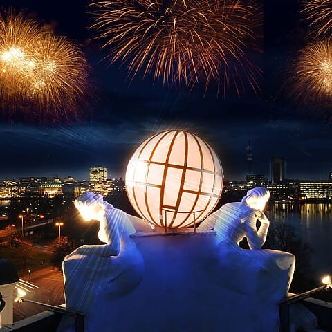 Feuerwerk über der Stadt mit leuchtender Skulptur und Nachtpanorama am Wasser.