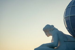 Skulptur einer sitzenden Frau mit Globus im Hintergrund bei Sonnenuntergang.