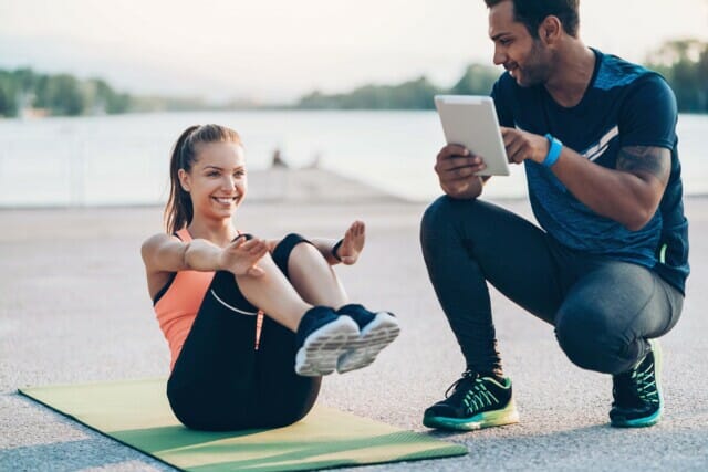 Junge Frau macht Fitnessübungen auf einer Matte, während ein Trainer sie anleitet.