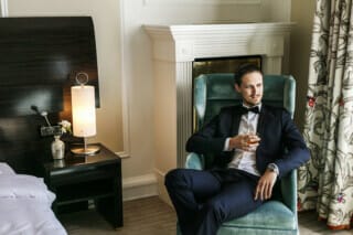 Elegant gekleideter Mann in einem modernen Hotelzimmer mit Getränk in der Hand.