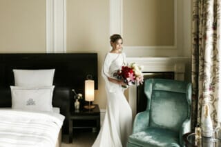 Braut im eleganten Kleid mit Blumenstrauß in einem stilvollen Hotelzimmer.