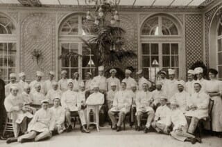 Historisches Gruppenfoto von Köchen in einer eleganten Umgebung mit Pflanzen und Fenstern.