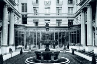 Innenhof mit Springbrunnen und Säulen, umgeben von eleganten Glaswänden und Sitzbereichen.