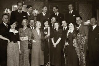 Gruppenfoto von elegant gekleideten Menschen in einem historischen Raum, um ein Mikrofon versammelt.