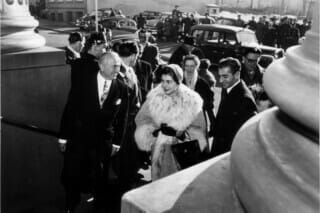 Historische Schwarz-Weiß-Aufnahme einer eleganten Frau mit Pelzmantel, umgeben von Menschen.