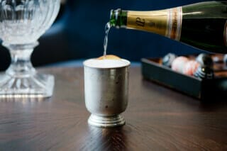 Sekt wird in einen eleganten Becher mit einer Orangenscheibe gegossen, auf einem Holztisch.