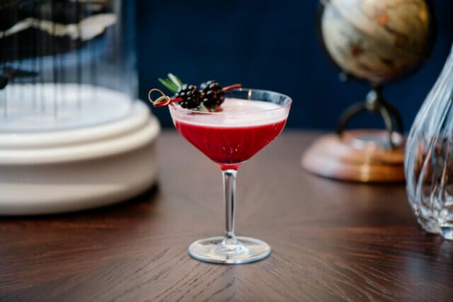 Eleganter Cocktail in einem Glas, rot mit Beeren und Rosmarin, auf einem Holztisch.