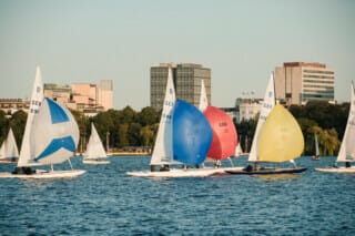 Hotel Atlantic Segelboote mit bunten Segeln auf einem See vor einer Stadtlandschaft bei Sonnenuntergang.