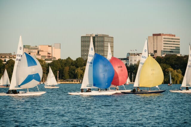 Segelboote mit bunten Segeln auf einem See vor einer Stadtlandschaft bei Sonnenuntergang.