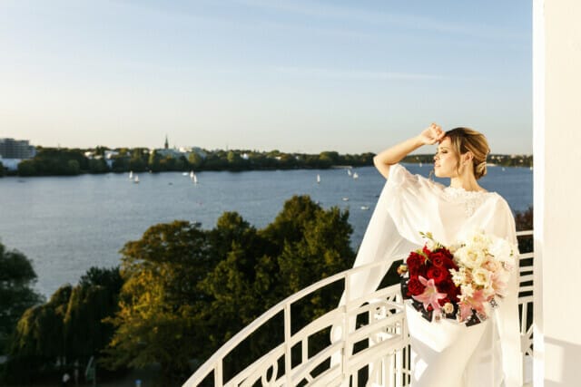 Braut in eleganter Robe mit Blumenstrauß, Blick auf den See und die Stadt im Hintergrund.