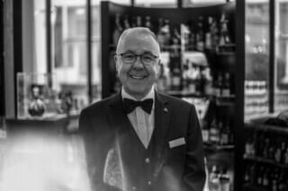 Hotel Atlantic Lächelnder Barkeeper in eleganter Schwarz-Weiß-Kleidung vor einer Bar mit Spirituosen.