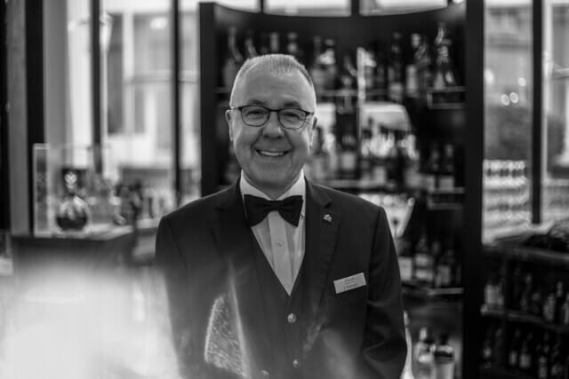 Lächelnder Barkeeper in eleganter Schwarz-Weiß-Kleidung vor einer Bar mit Spirituosen.