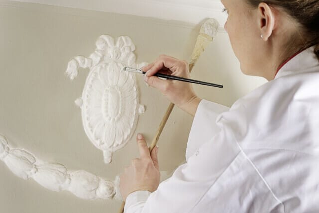 Frau malt kunstvolle Stuckverzierungen an einer Wand mit einem Pinsel und Holzstab.
