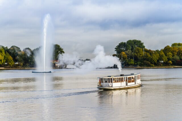 Dampfschiff fährt auf ruhigem Wasser, im Hintergrund ein Wasserstrahl und grüne Bäume.