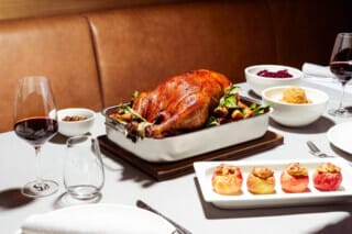 Cook in white jacket holding a roast duck on a serving tray with side dishes.