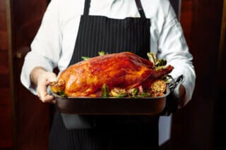 Cook in white jacket holding a roast duck on a serving tray with side dishes.