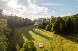 Landschaft mit Hotel im Hintergrund, umgeben von Bäumen und einer Wiese im Sonnenlicht.