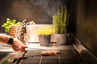 Ein Koch wendet ein Steak auf dem Grill, umgeben von frischen Kräutern und Küchengeräten.