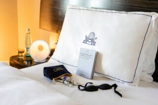 Elegantes Hotelzimmer mit Kissen, Schlafmaske, Pflegeprodukten und Buch auf dem Bett.