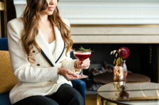Frau mit einem Cocktail in der Hand, elegant gekleidet, sitzt vor einem Kamin.