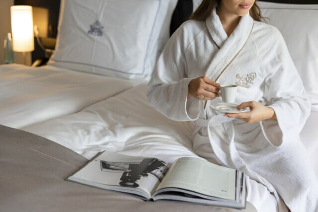 Frau in Bademantel mit Tasse Kaffee, entspannt auf einem Bett mit Zeitschrift.
