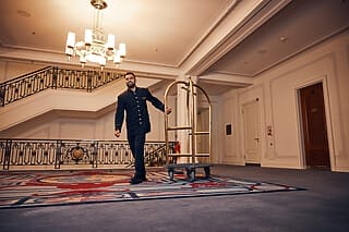 Hotelmitarbeiter mit Gepäckwagen in eleganter Lobby mit Treppe und kunstvollem Teppich.
