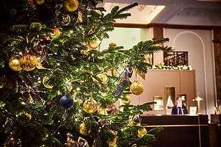 Weihnachtsbaum mit goldenen und blauen Kugeln, Lichtern und festlicher Dekoration im Hintergrund.