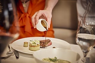 Eine Person gießen Sauce über ein kunstvoll angerichtetes Steakgericht mit Beilagen.