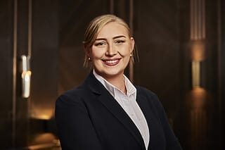 Professionelle Frau mit blonden Haaren in Anzug, lächelnd vor elegantem Hintergrund.