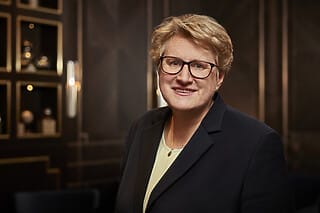 Professionelle Frau mit Brille und blauer Jacke vor elegantem Hintergrund.