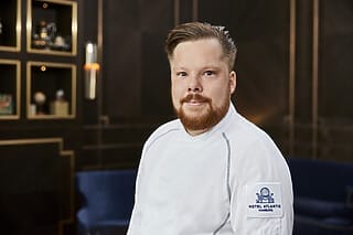 Koch des Hotel Atlantic Hamburg in weißer Uniform, lächelnd vor elegantem Hintergrund.