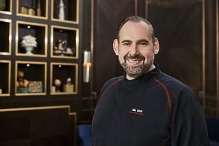 Koch Nils Uhlott in schwarzer Kochjacke vor dekorativem Hintergrund in einem Restaurant.
