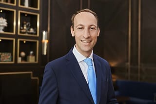 Lächelnder Mann in Anzug mit blauer Krawatte vor elegantem Hintergrund.