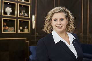 Professional woman with curly hair, smiling in front of an elegant background with shelves.