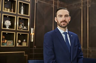 Mann im Anzug vor einer eleganten Wand mit Vitrinen und dekorativen Objekten.