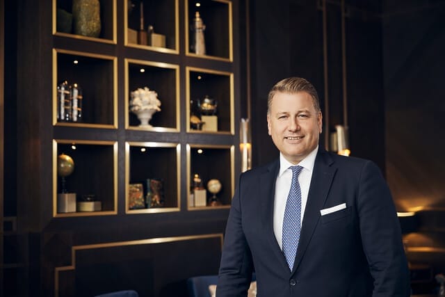 Geschäftsmann in Anzug lächelt vor einer eleganten Wand mit dekorativen Regalen.