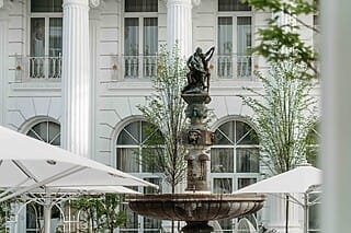 Hotel Atlantic Brunnen mit Statue vor einem eleganten Gebäude mit Säulen und Bäumen im Hintergrund.