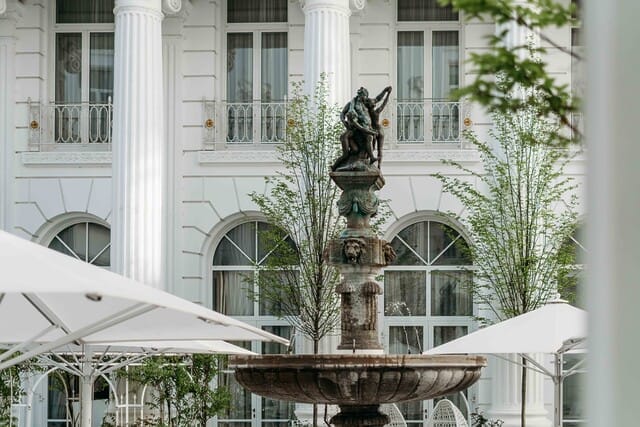 Brunnen mit Statue vor einem eleganten Gebäude mit Säulen und Bäumen im Hintergrund.