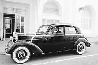 Hochzeitsauto in Schwarz mit Brautpaar, dekoriert mit Schleifen, vor einem eleganten Gebäude.