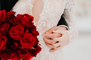 Braut mit tiefem Ausschnitt in Spitzenkleid, hält roten Rosenstrauß und Hand des Bräutigams.