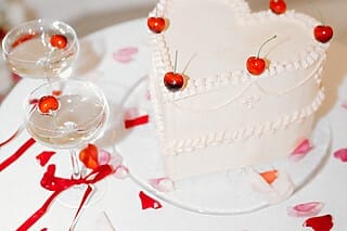 Herzförmiger Kuchen mit Kirschen, umgeben von Sektgläsern und Blütenblättern auf Tisch.
