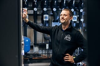 Techniker in schwarzem Pullover steht vor Maschinen, lächelt und zeigt auf einen Schalter.
