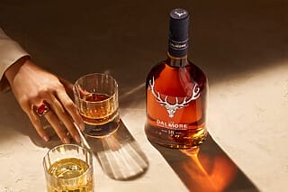 Flasche Dalmore 18 Jahre Whisky mit Gläsern und Hand auf einem eleganten Tisch.
