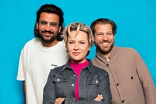 Drei Menschen posieren vor einem blauen Hintergrund, lächelnd und in modischer Kleidung.