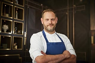 Koch in weißer Uniform mit blauer Schürze, lächelt selbstbewusst in modernem Restaurant.