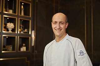Koch im Hotel Atlantic Hamburg lächelt vor einer eleganten Kulisse mit dekorativen Regalen.