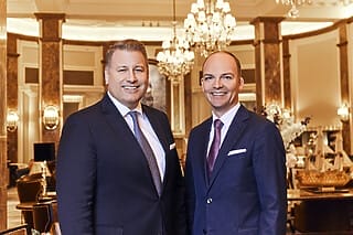 Zwei lächelnde Männer in Anzügen stehen in einem eleganten Hotel-Lobby.