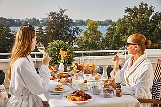 Zwei Frauen im Bademantel genießen ein Frühstück auf dem Balkon mit Blick auf den See.